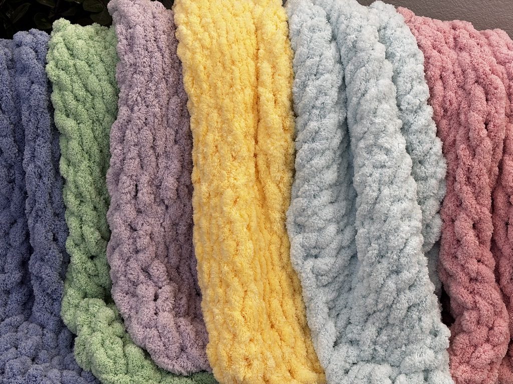 A row of pastel handmade baby blankets hanging side by side in blue, green, lavender, yellow, sky and pink.