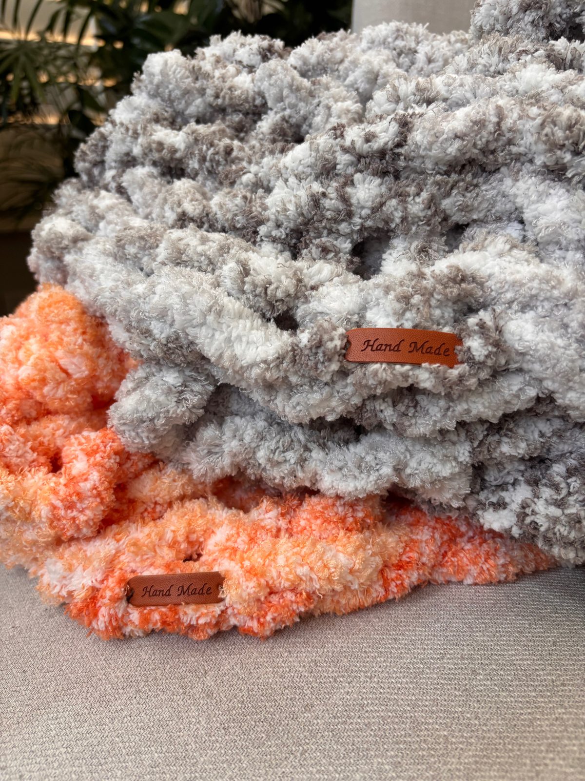 A grey baby blanket and a peach baby blanket folded together, each with a leather Hand Made tag.