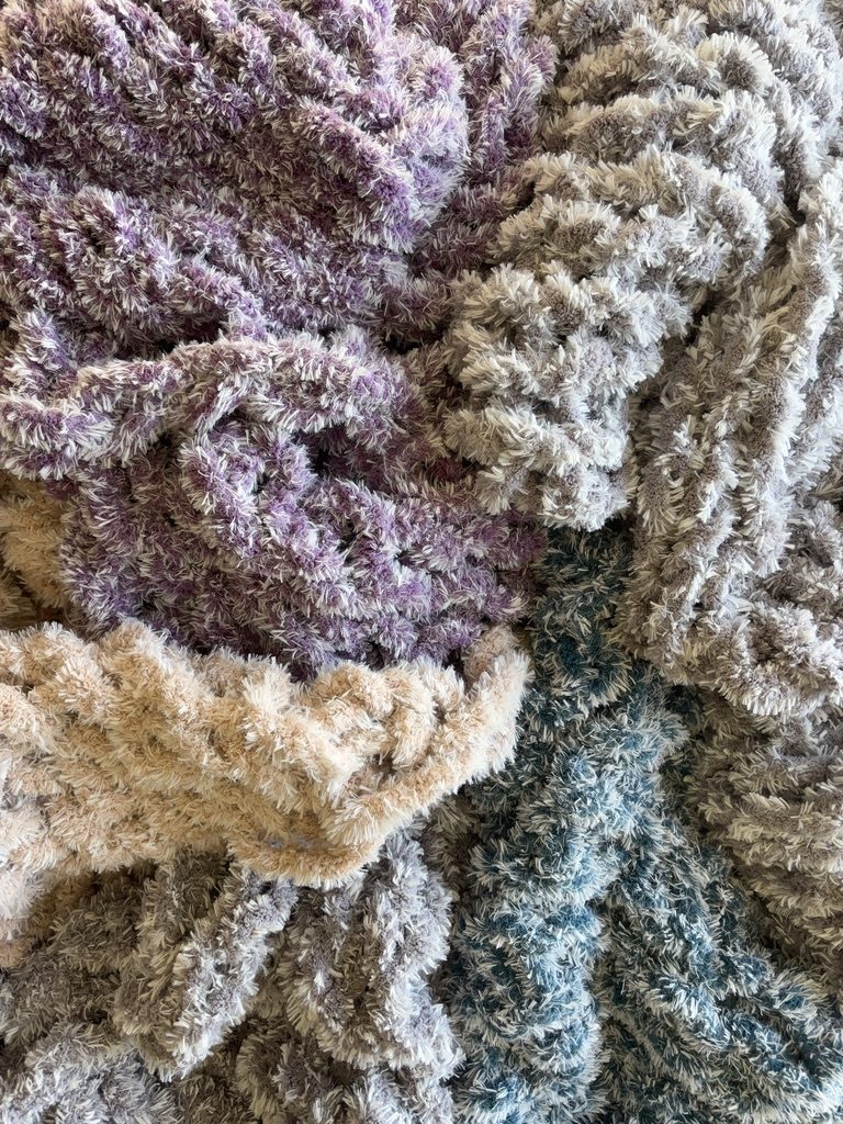 A close-up of textured chunky knits in lavender, taupe, cream and teal.